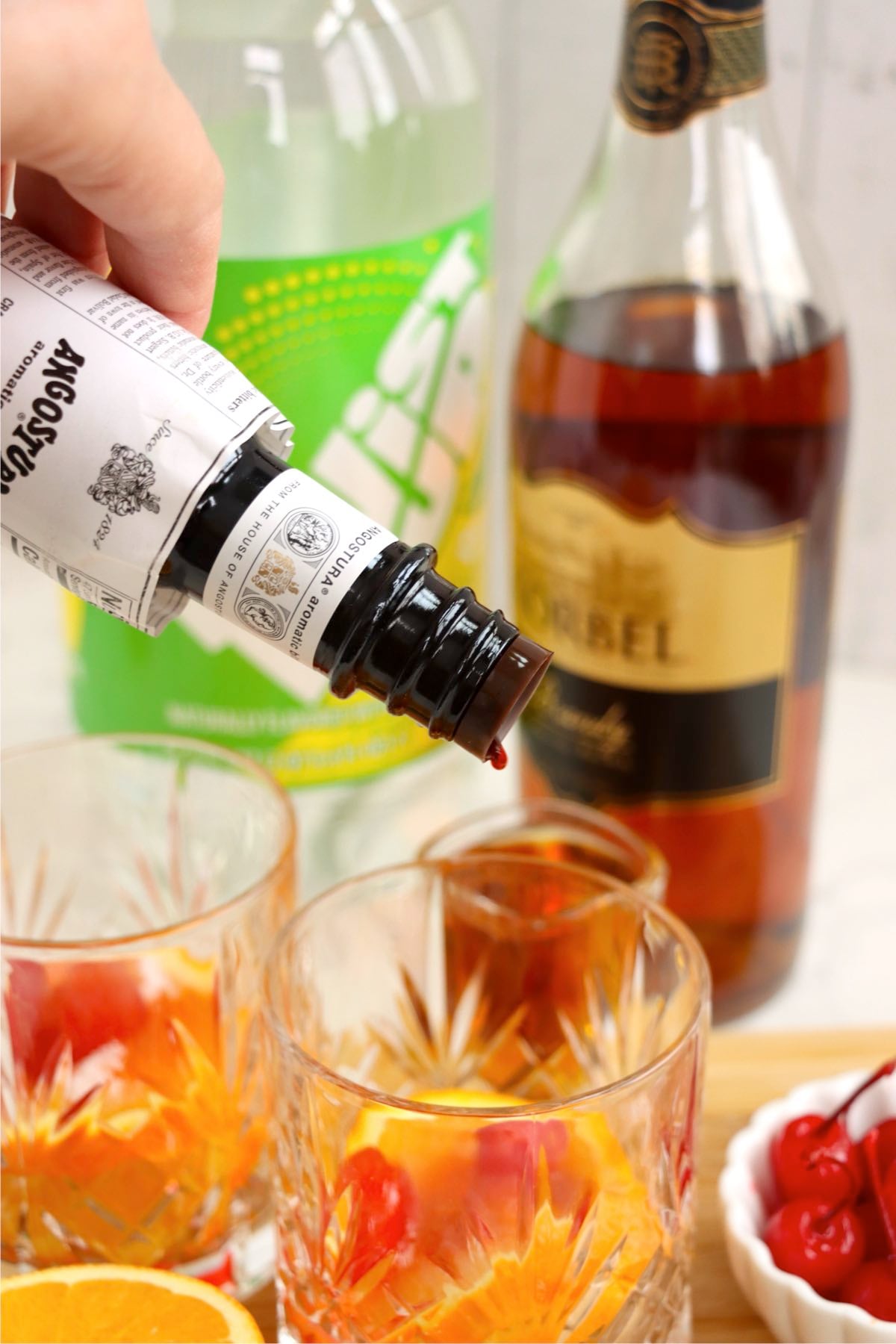 Angostura bitters being poured into an Old Fashioned glass with ice, with brandy and lemon-lime soda in the background.
