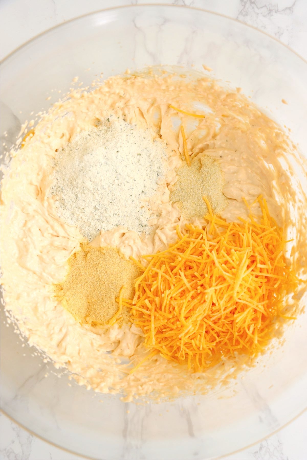 A bowl of creamy cheese mixture topped with shredded cheddar cheese, ranch seasoning, garlic powder, and onion powder before being stirred.