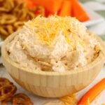 A bowl of cheese spread placed in front of a snack tray with pretzels, celery, and carrots in the background.