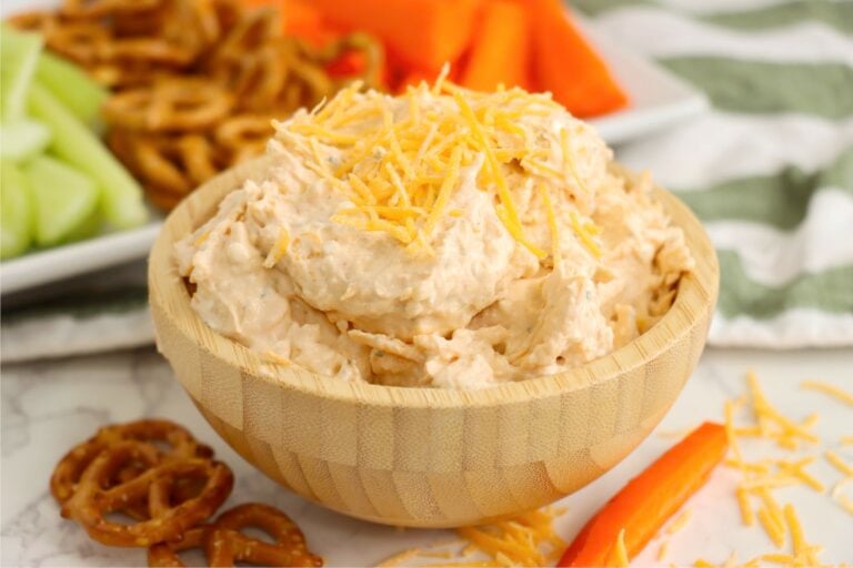 A bowl of cheese spread placed in front of a snack tray with pretzels, celery, and carrots in the background.