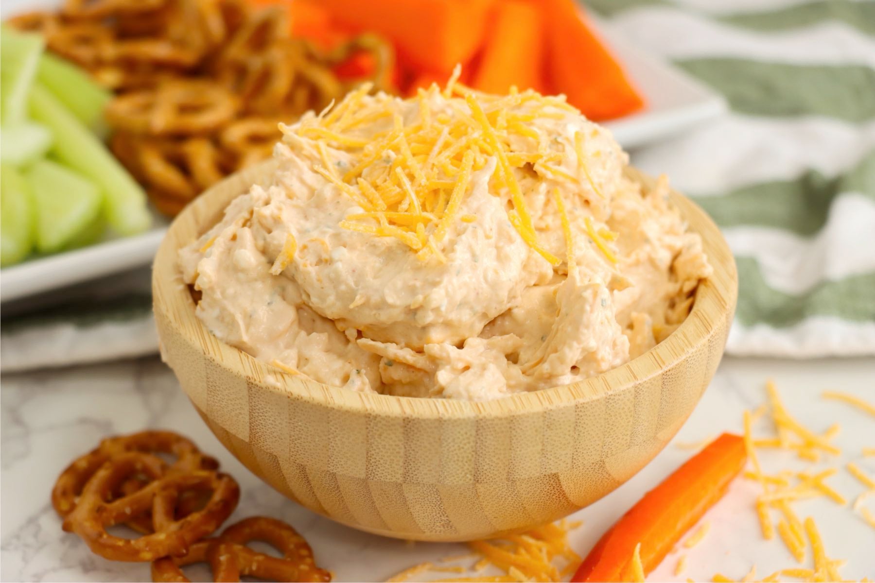 A bowl of cheese spread placed in front of a snack tray with pretzels, celery, and carrots in the background.