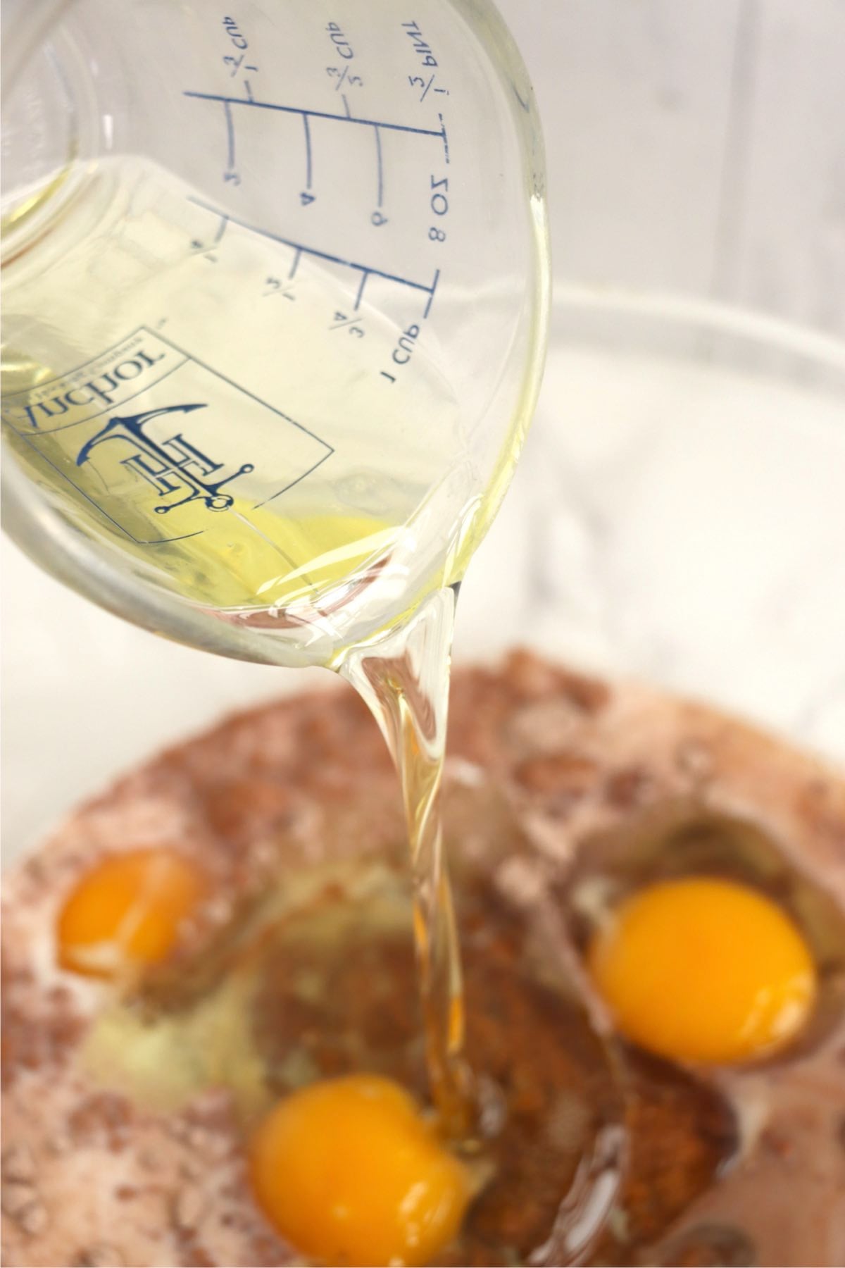 Vegetable oil being poured into a bowl of chocolate cake batter with eggs.