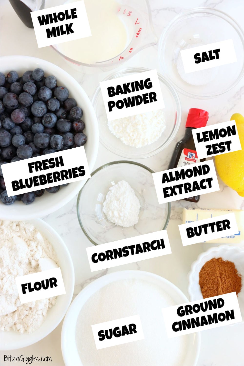 Blueberries, flour, sugar, milk, lemon zest, cinnamon and more ingredients laid out on a white marble table.