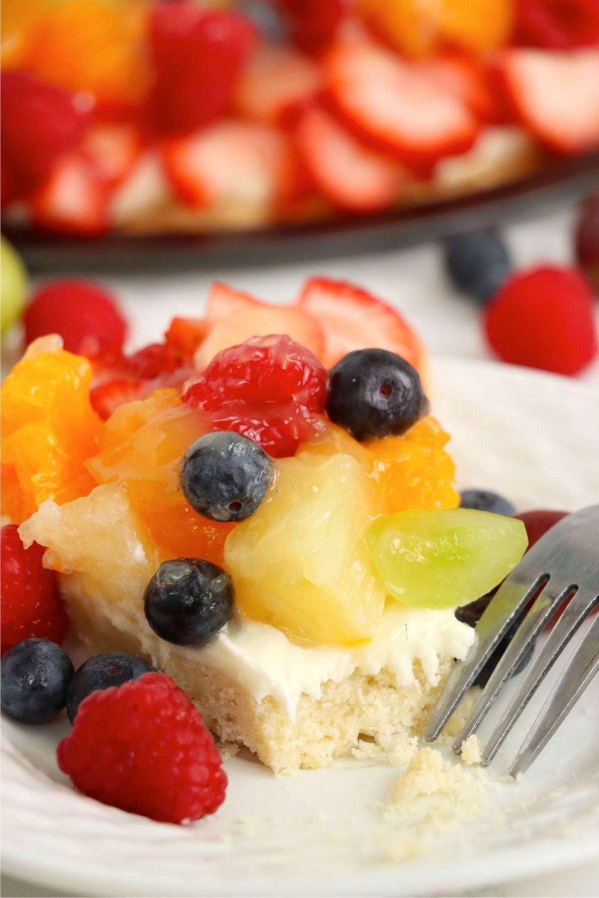A half eaten slice of fruit tart on a plate topped with strawberries, blueberries, pineapple, and grapes.