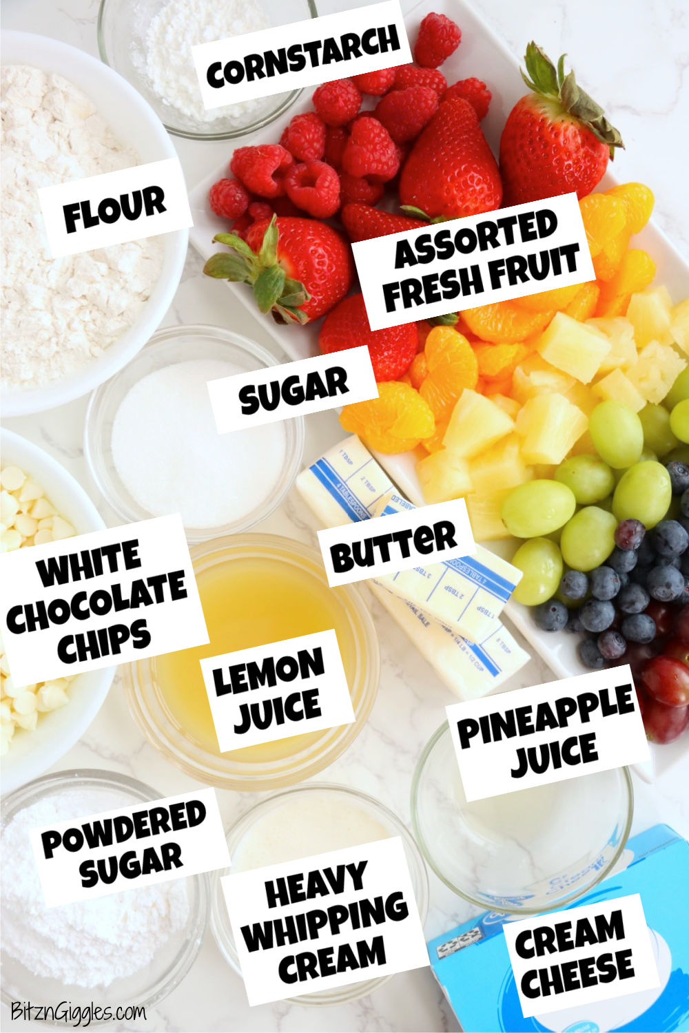 Assorted ingredients for a fruit tart including fresh fruit, flour, butter, cream cheese, white chocolate chips, sugar, and juices arranged on a countertop.