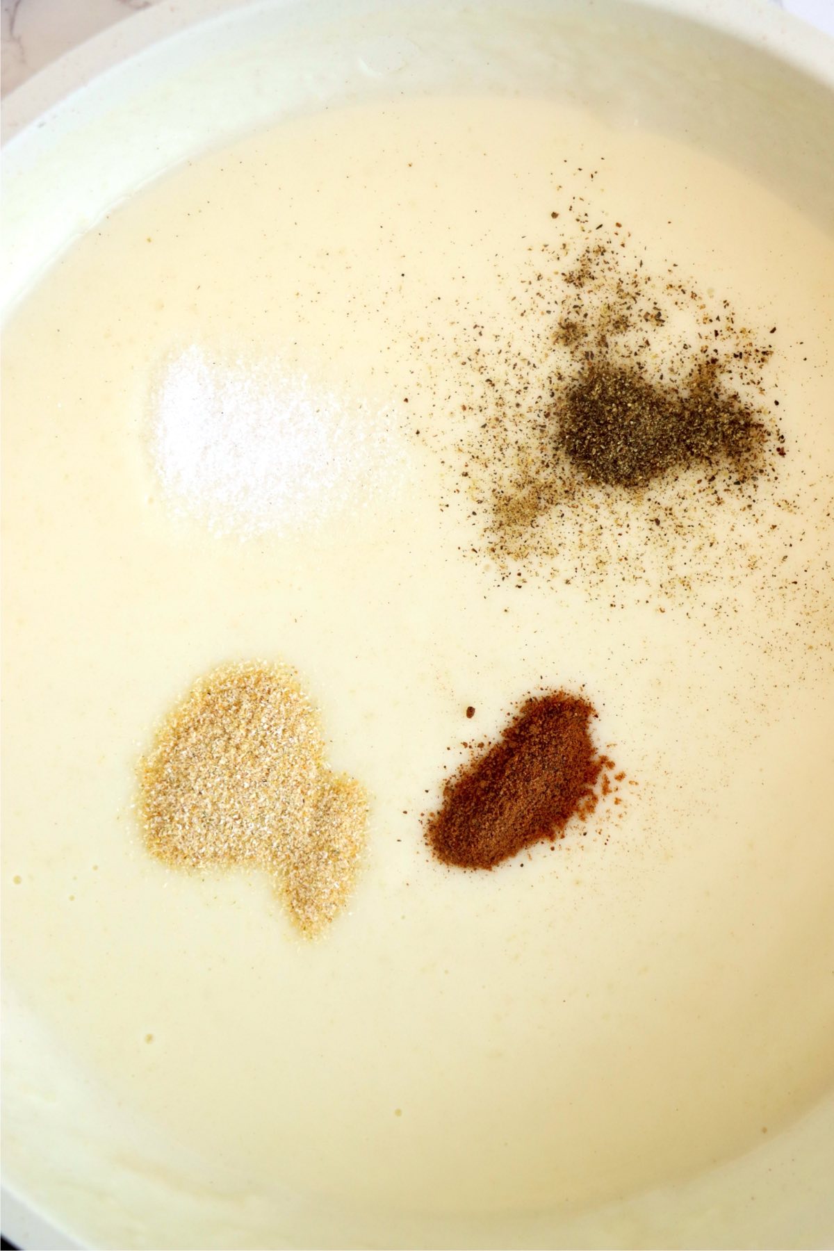 Cream sauce with salt, pepper, onion powder, and nutmeg added in a saucepan.