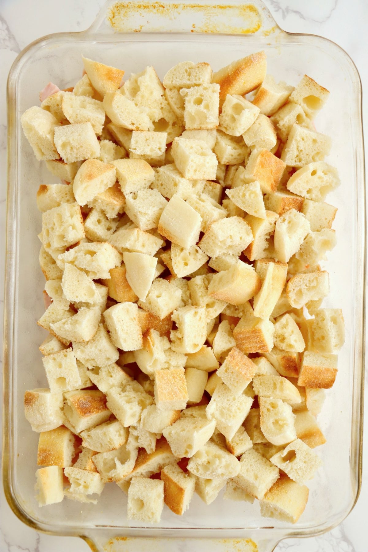 Cubed English muffins layered over chopped Canadian bacon in a casserole dish.