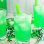 Three glasses of bright green lemonade with ice and green straws surrounded by green shamrock decorations.