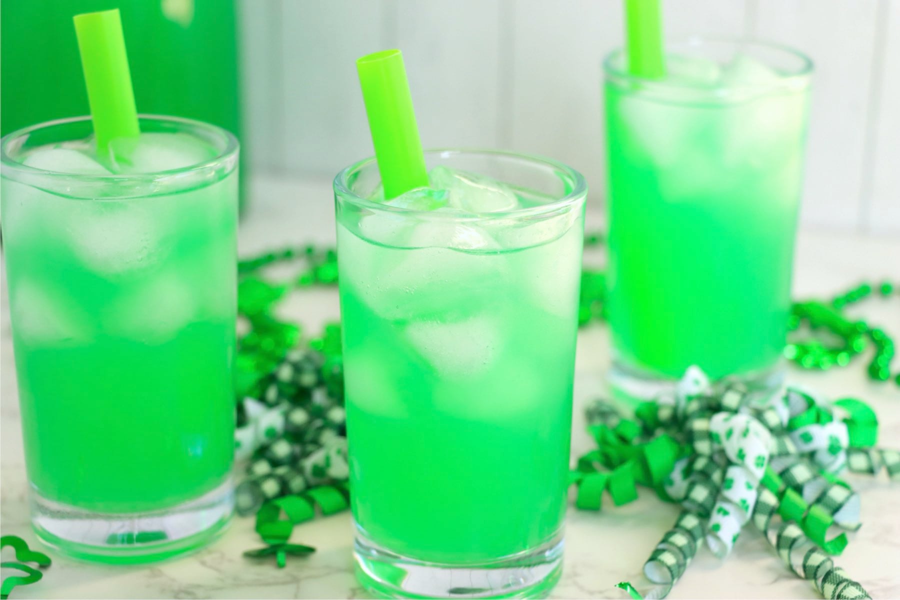 Three glasses of bright green lemonade with ice and green straws surrounded by green shamrock decorations.