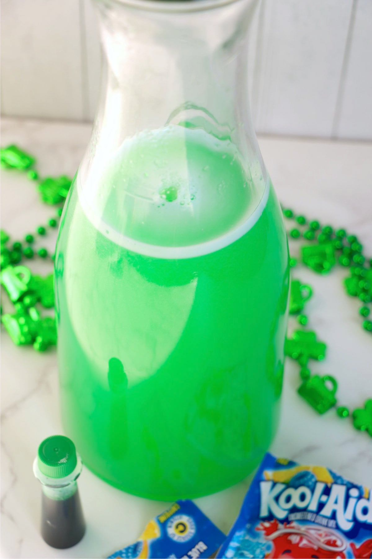 Glass pitcher filled with bright green lemonade sitting on a countertop with green shamrock decorations nearby.