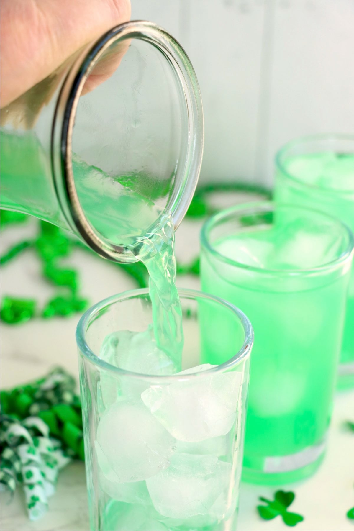 Green lemonade being poured from a pitcher into glasses filled with ice.