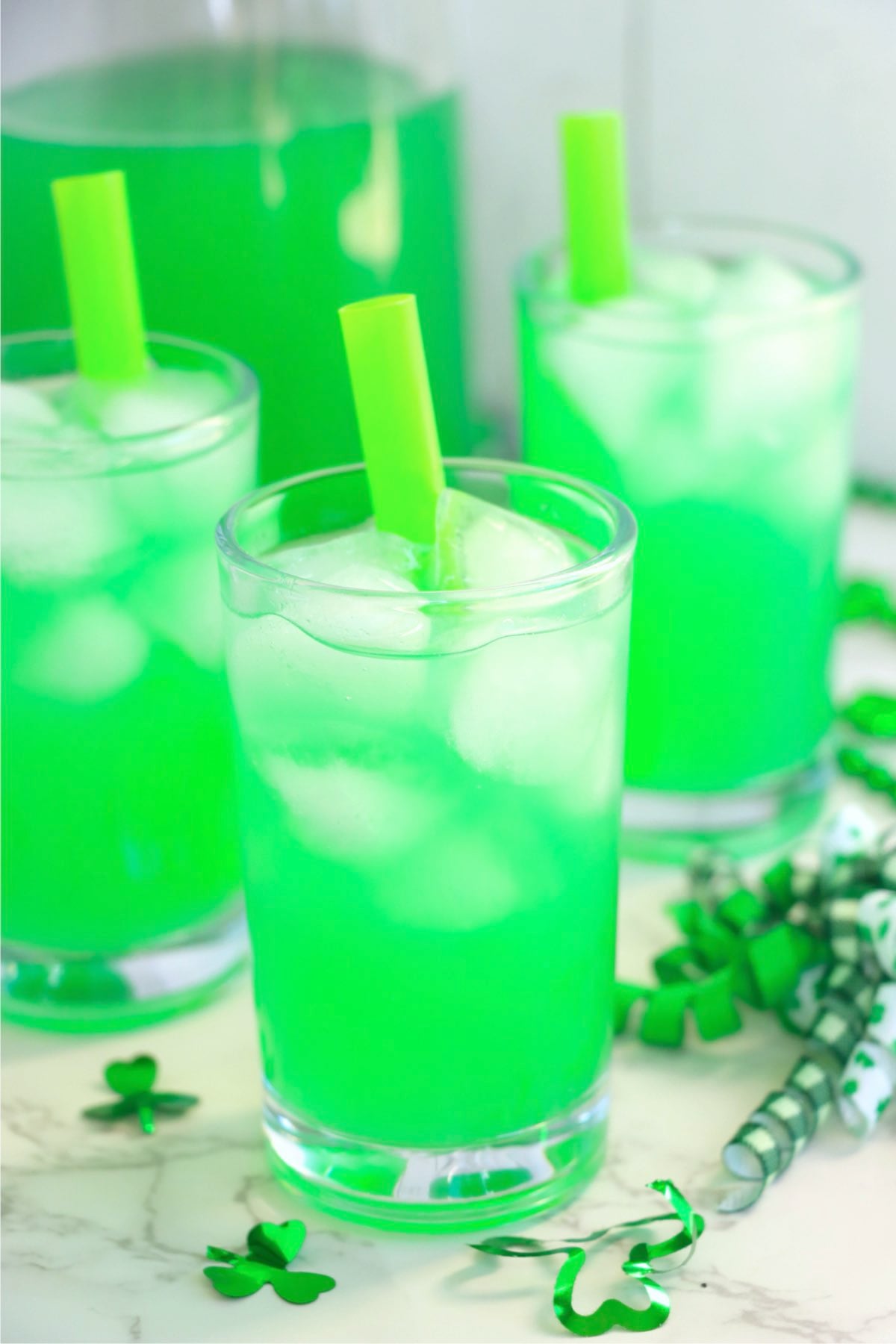Three glasses of green lemonade with ice and green straws with a pitcher of the drink in the background.