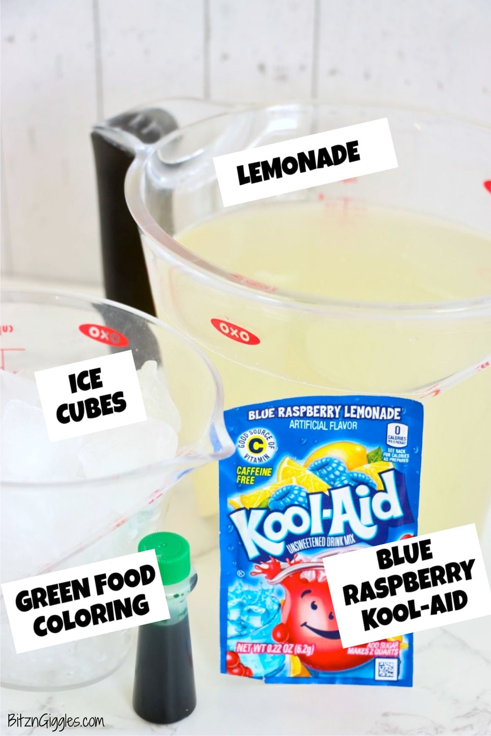 Measuring cups with lemonade and ice cubes next to a packet of blue raspberry drink mix and a bottle of green food coloring.