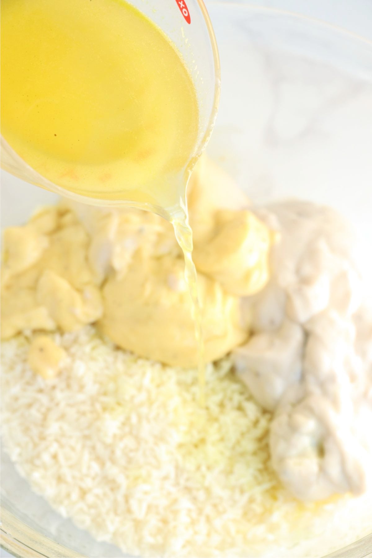 Chicken broth being poured over uncooked rice and condensed soups in a glass mixing bowl.