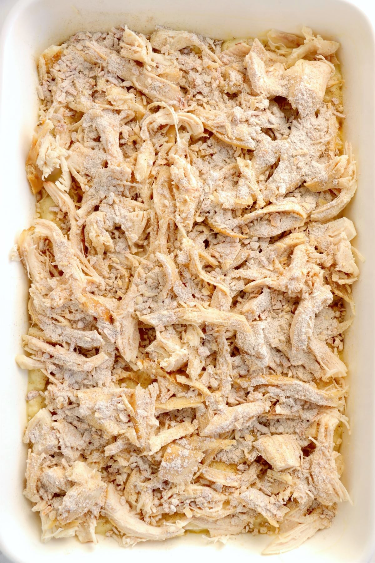 Shredded rotisserie chicken layered over creamy rice mixture in a white casserole dish before baking.