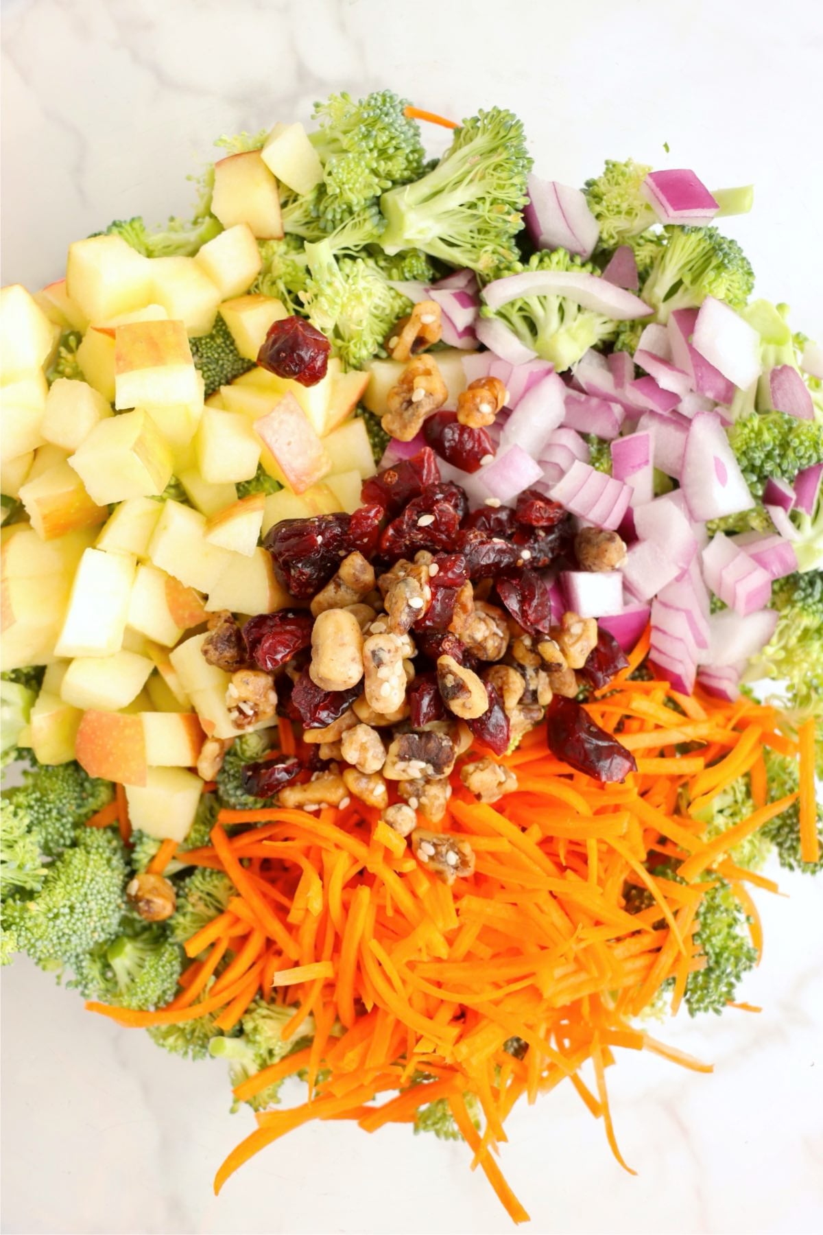 Fresh broccoli salad ingredients including apples, carrots, cranberries, walnuts, and red onion in a mixing bowl.