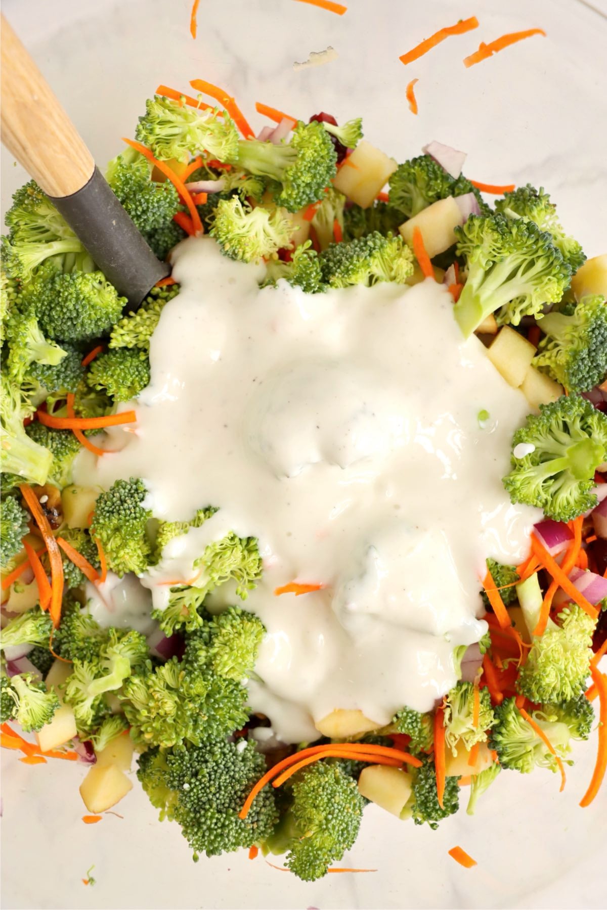 Creamy yogurt dressing poured over fresh broccoli, carrots, apples, and onions in a large bowl.