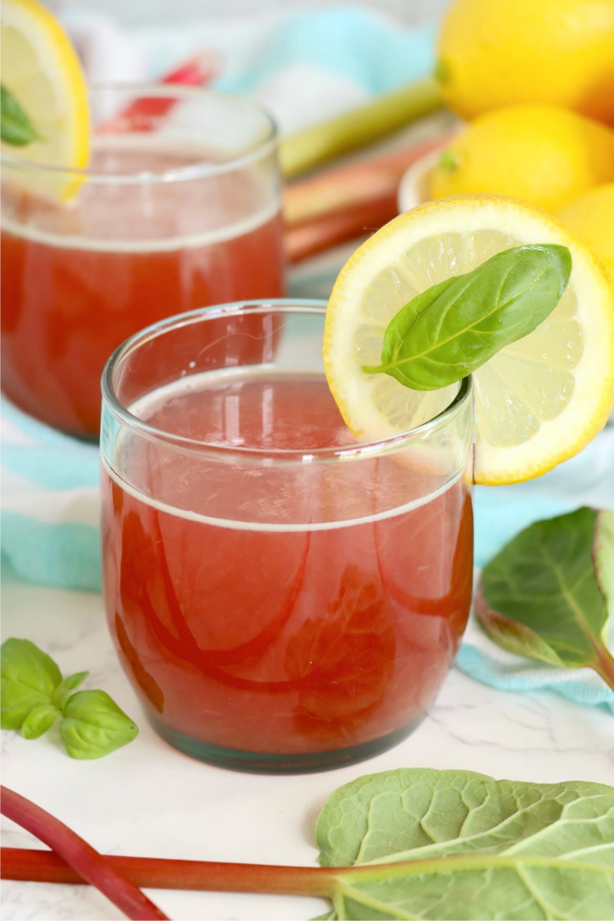 Rhubarb cordial garnished with a lemon slice and fresh basil leaf, surrounded by fresh rhubarb stalks and lemons.