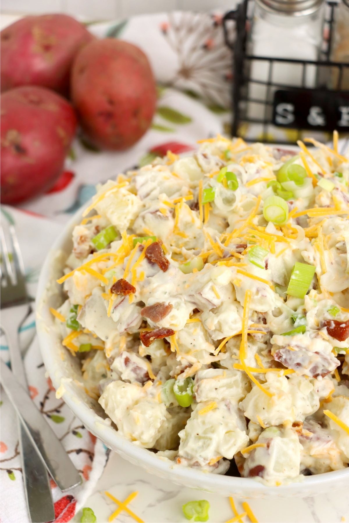 Creamy potato salad topped with shredded cheddar cheese, crispy bacon pieces and sliced green onions in a white serving bowl.