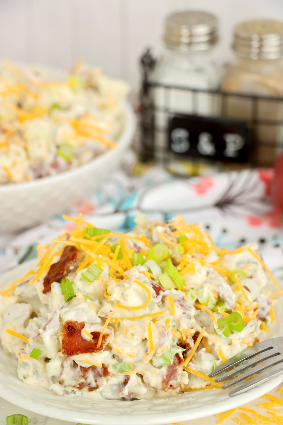 Creamy potato salad served on a white plate with cheddar cheese, bacon crumbles and sliced green onions.