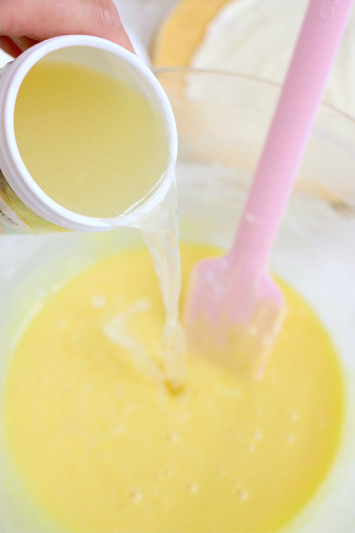 Lemonade concentrate being poured into creamy yellow mixture beside a pink spatula.