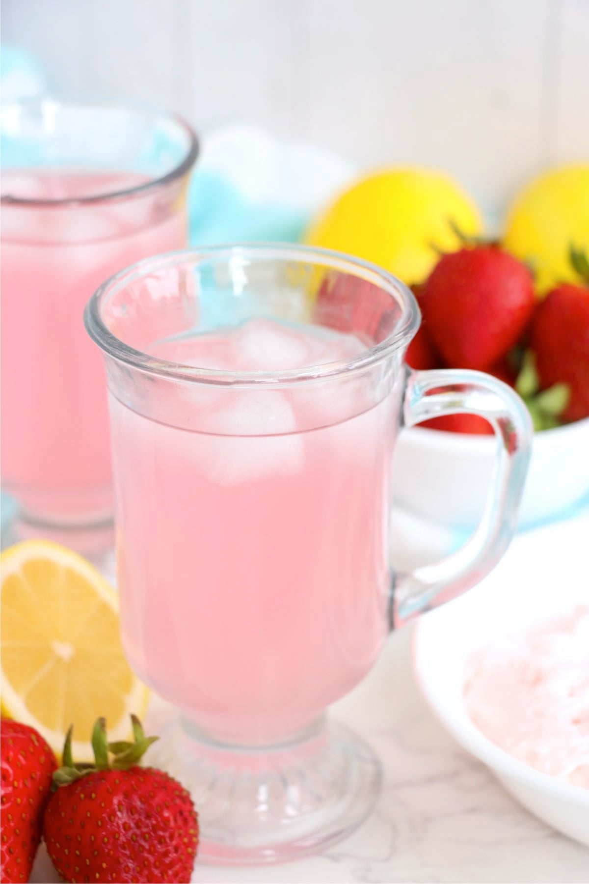 Glass of pink lemonade over ice with fresh lemons and strawberries nearby.