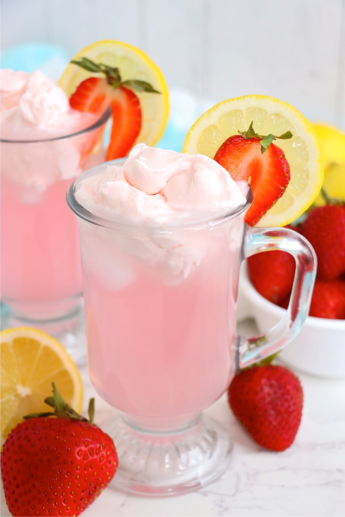 Two glasses of pink lemonade topped with whipped cream and fresh fruit garnishes.