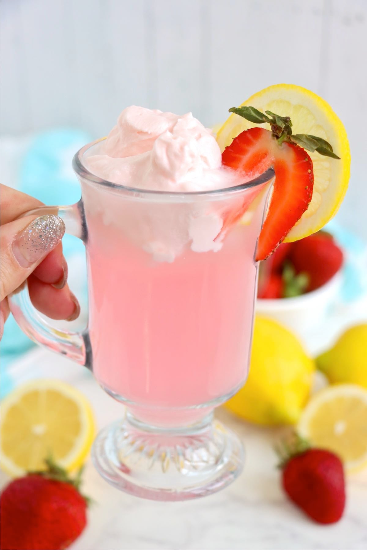 Hand holding whipped pink lemonade topped with whipped cream, lemon and strawberry.