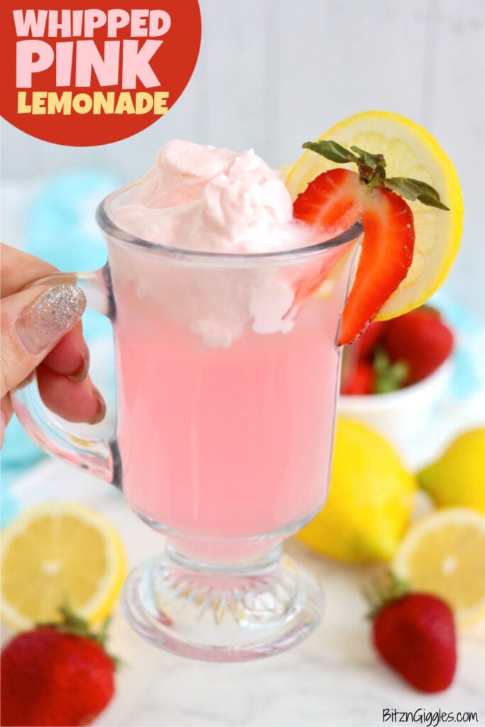 Hand holding a glass of pink lemonade topped with whipping cream.