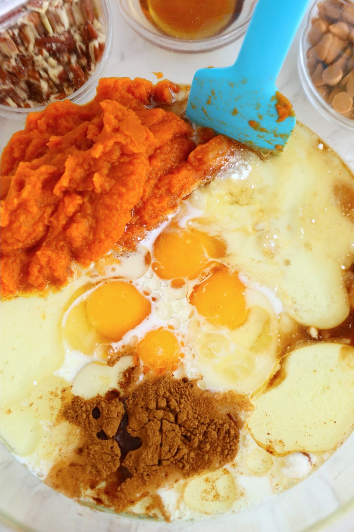 Pumpkin purée, eggs, milk, oil and brown spice sit unmixed in a glass bowl beside pecans and butterscotch chips.