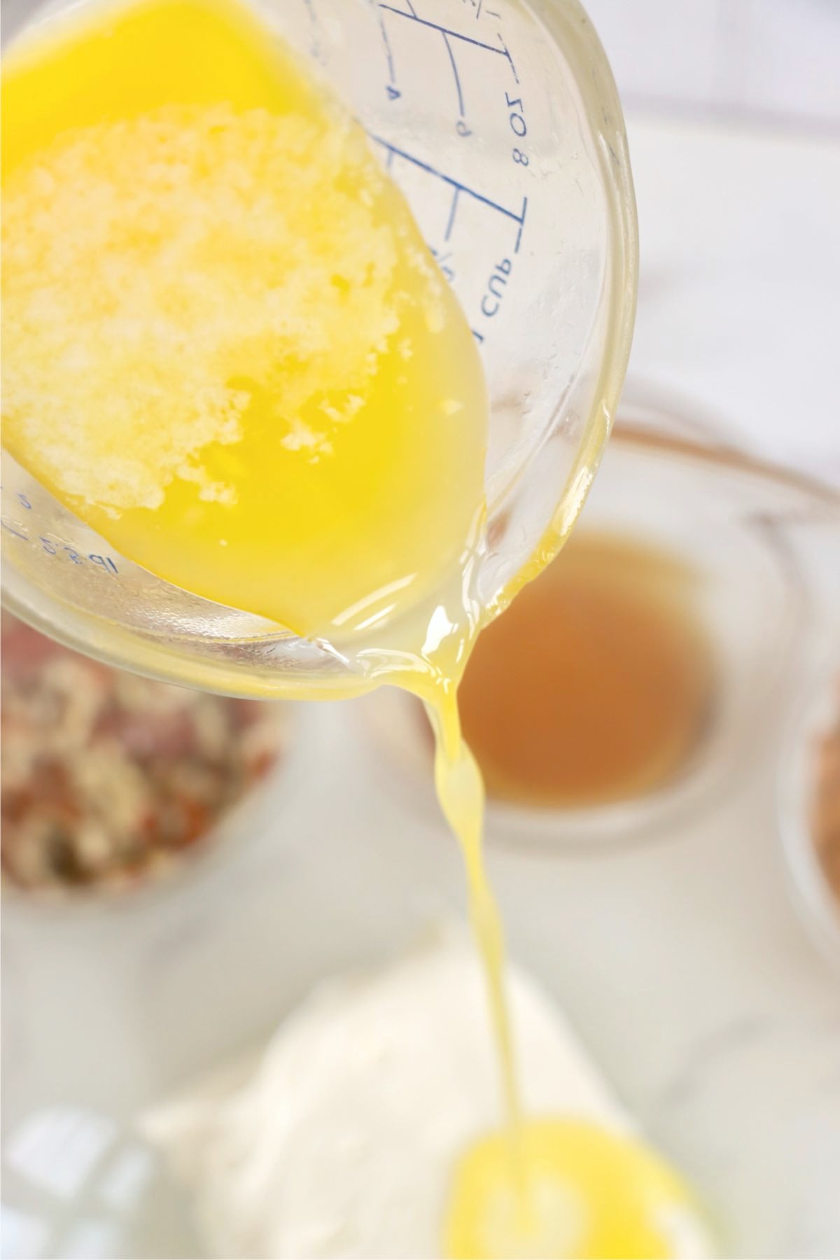 Melted butter pours from a glass measuring cup toward creamy white ingredients in a mixing bowl.
