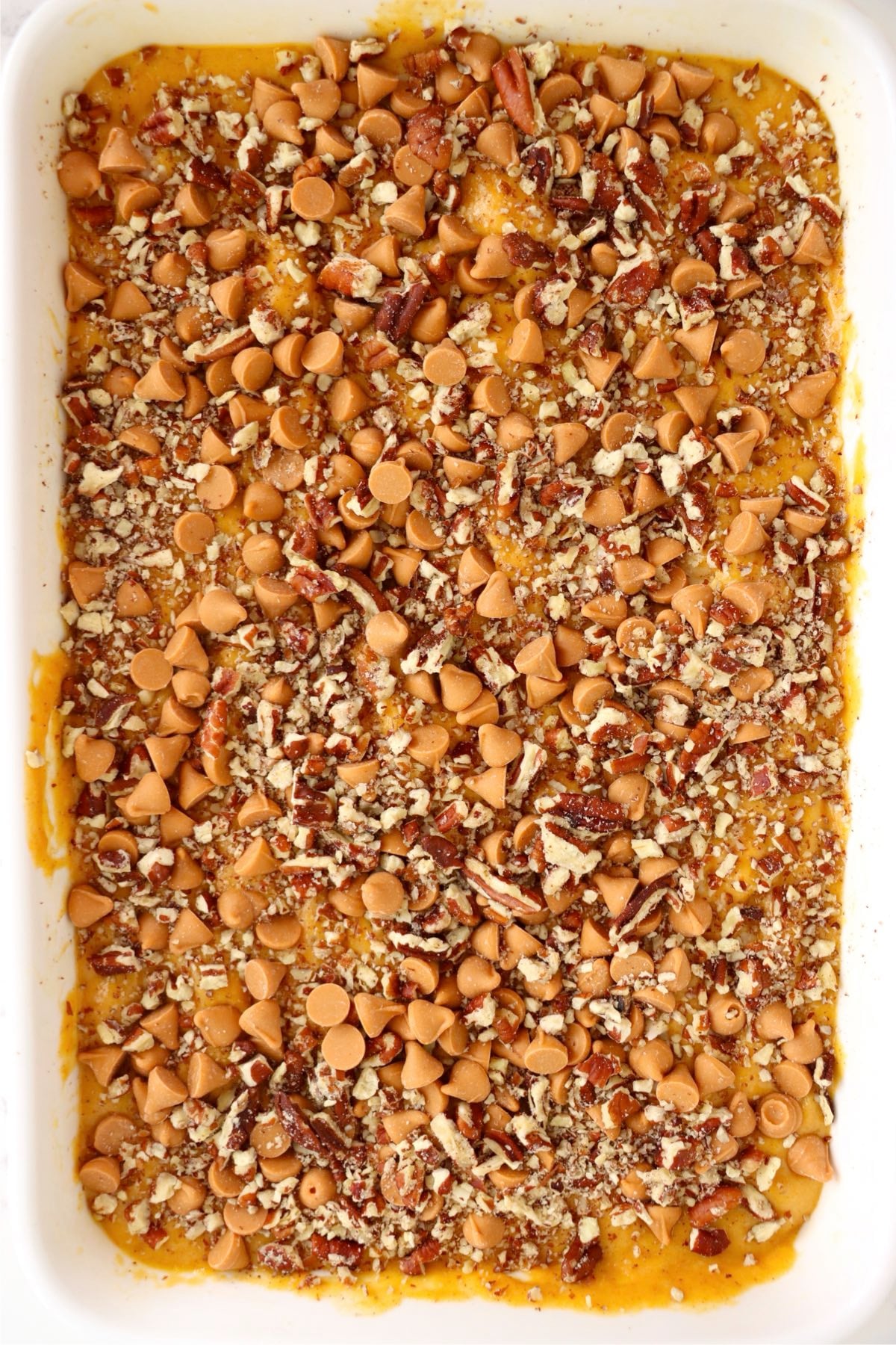 Orange cake batter fills a white baking dish beneath chopped pecans and scattered butterscotch chips.