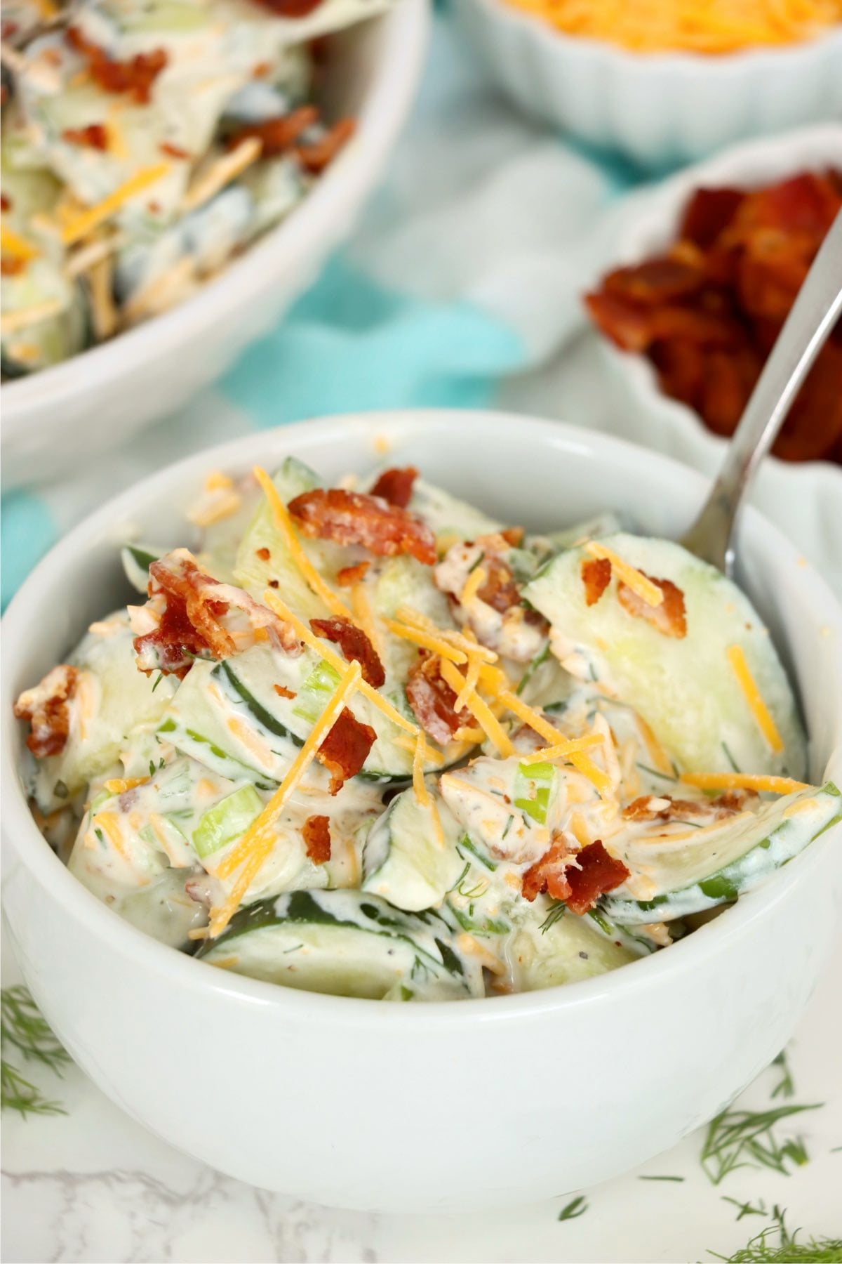 Small serving bowl of creamy cucumber slices with bacon and cheddar surrounded by extra toppings.