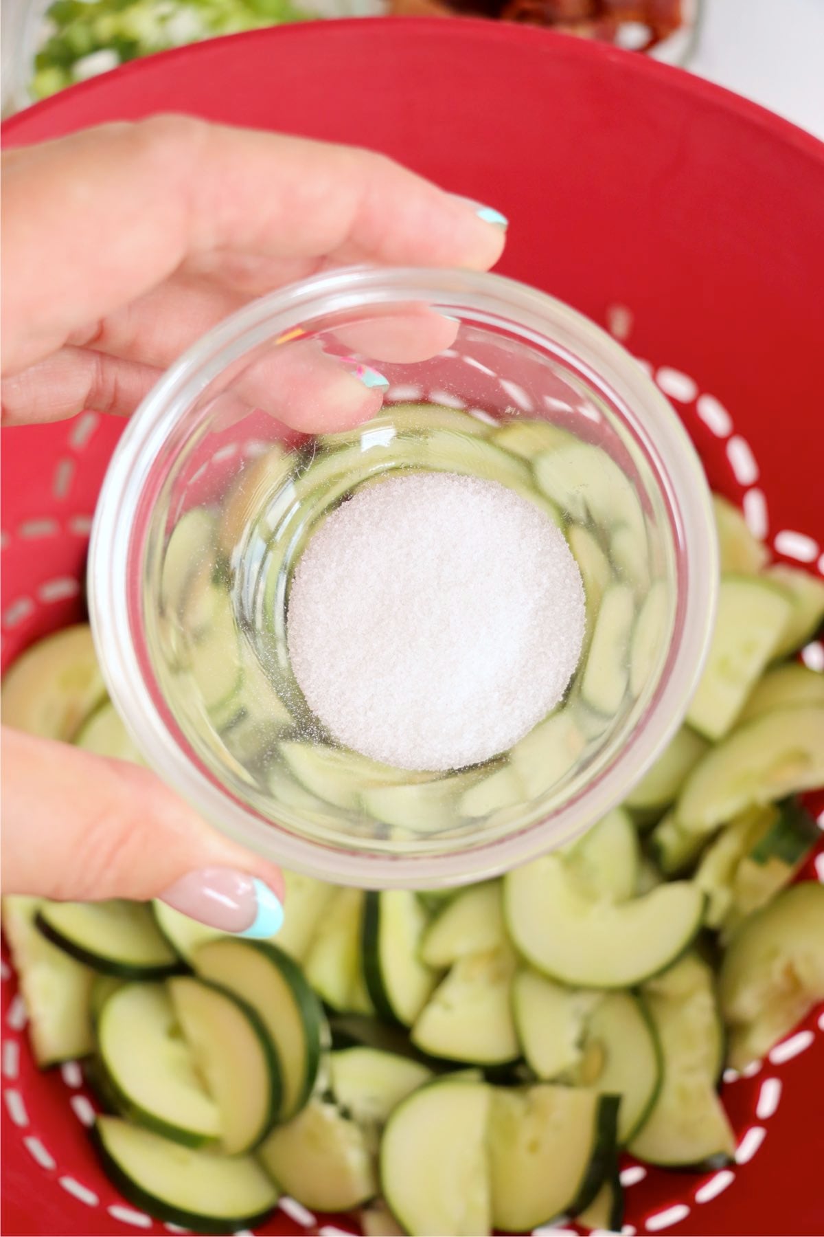 Kosher salt held over sliced cucumber half-moons draining in a bright red colander.