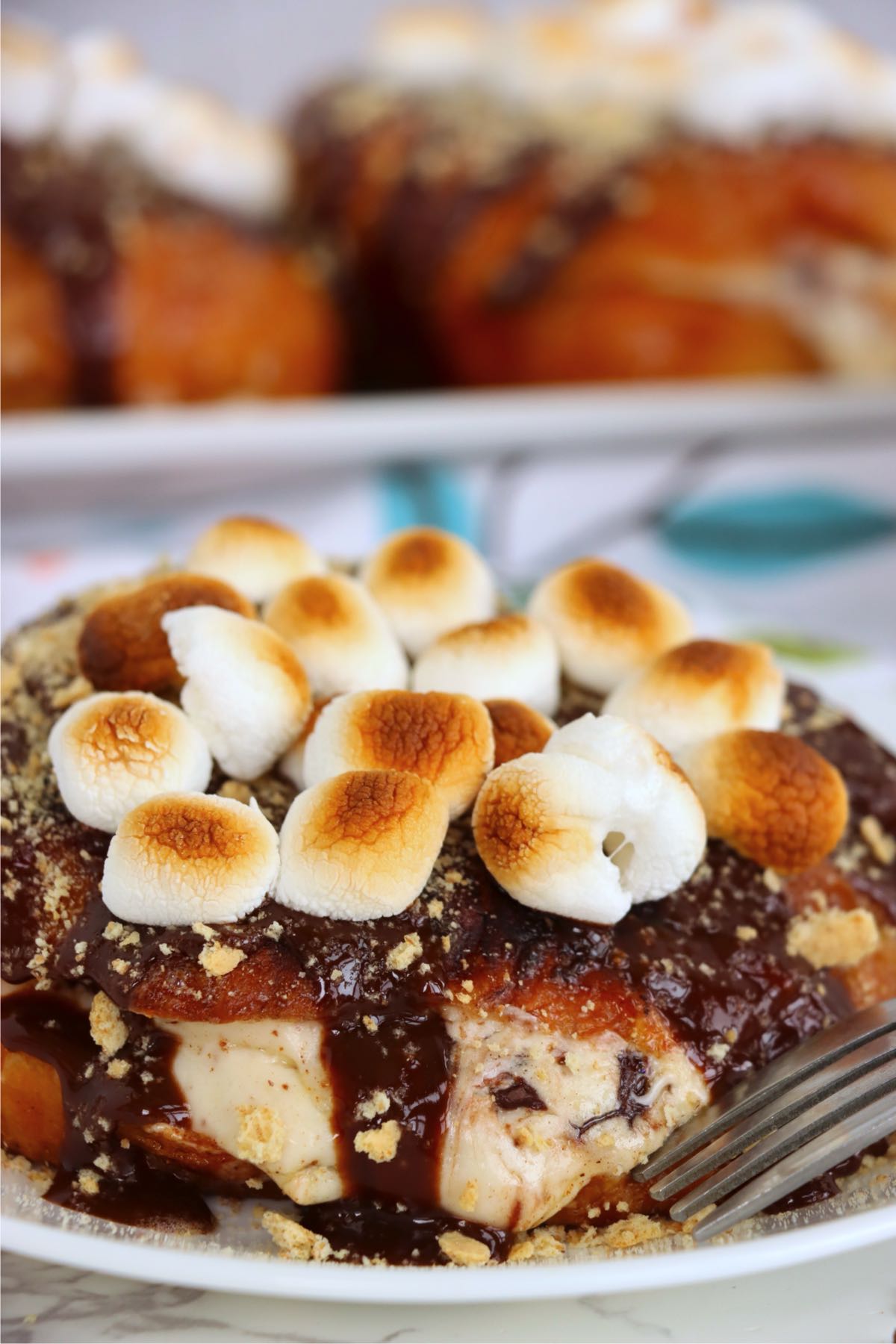 Close-up of toasted mini marshmallows piled over glossy chocolate sauce and graham cracker crumbs on a croissant.
