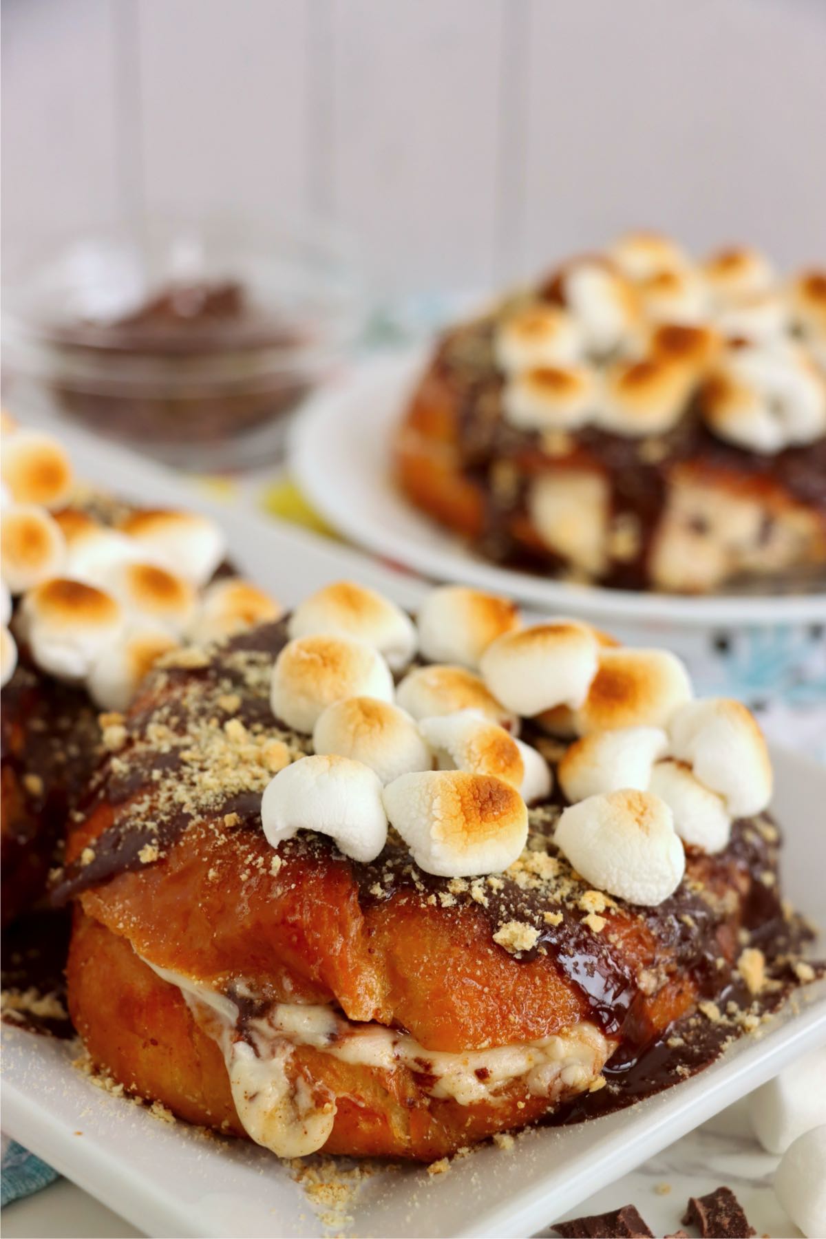 Three chocolate-drizzled stuffed croissants topped with toasted marshmallows and graham crumbs on a white serving platter.