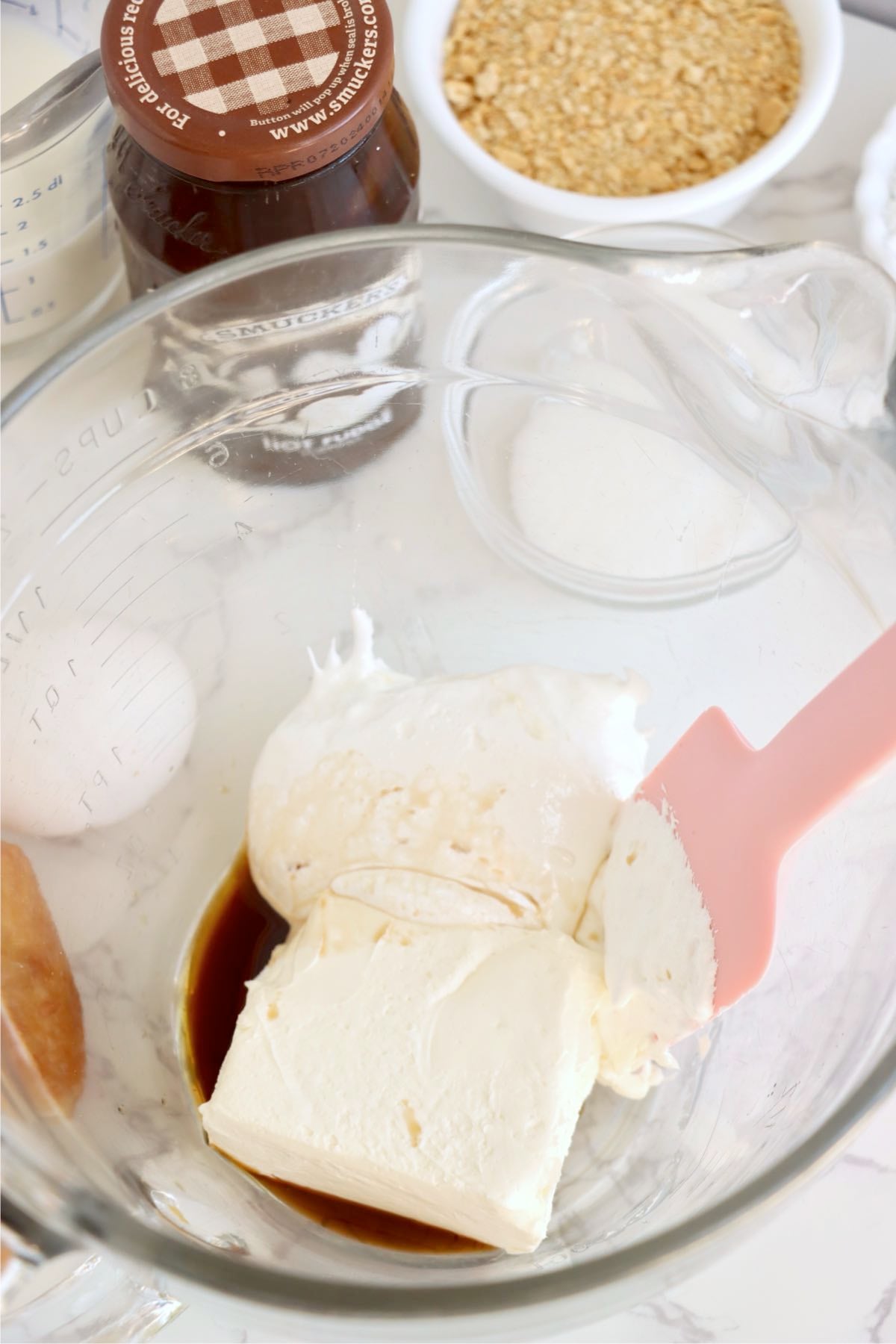 Softened cream cheese and vanilla extract in a glass mixing bowl beside milk, hot fudge and graham crumbs.