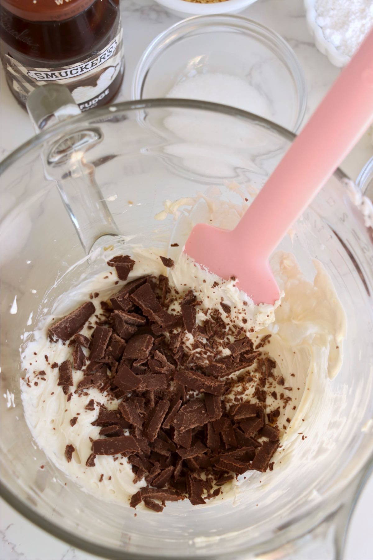 Chopped milk chocolate scattered over creamy marshmallow filling in a glass bowl with a pink spatula.