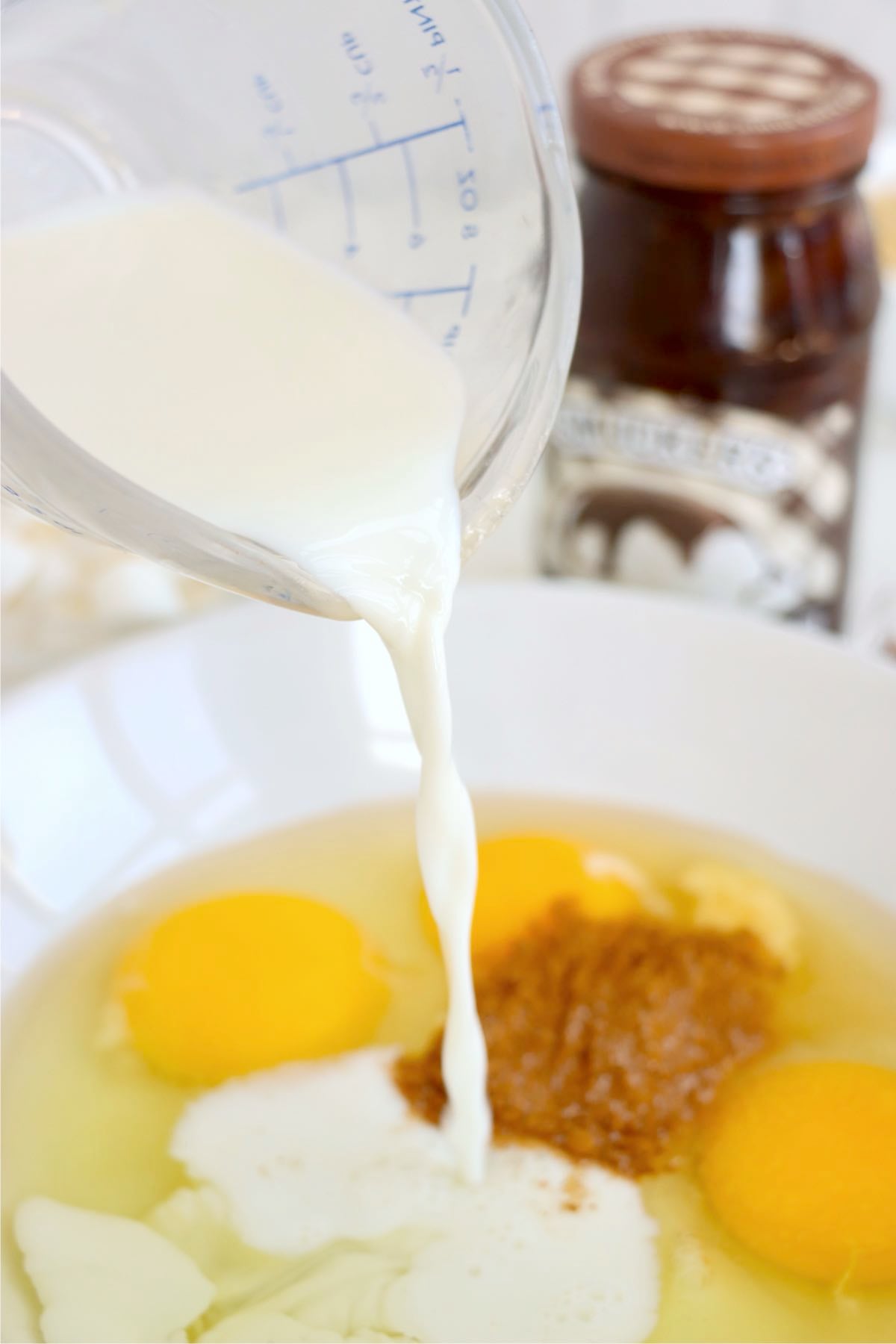 Milk pouring into a bowl of eggs, cinnamon and cream beside a jar of hot fudge.