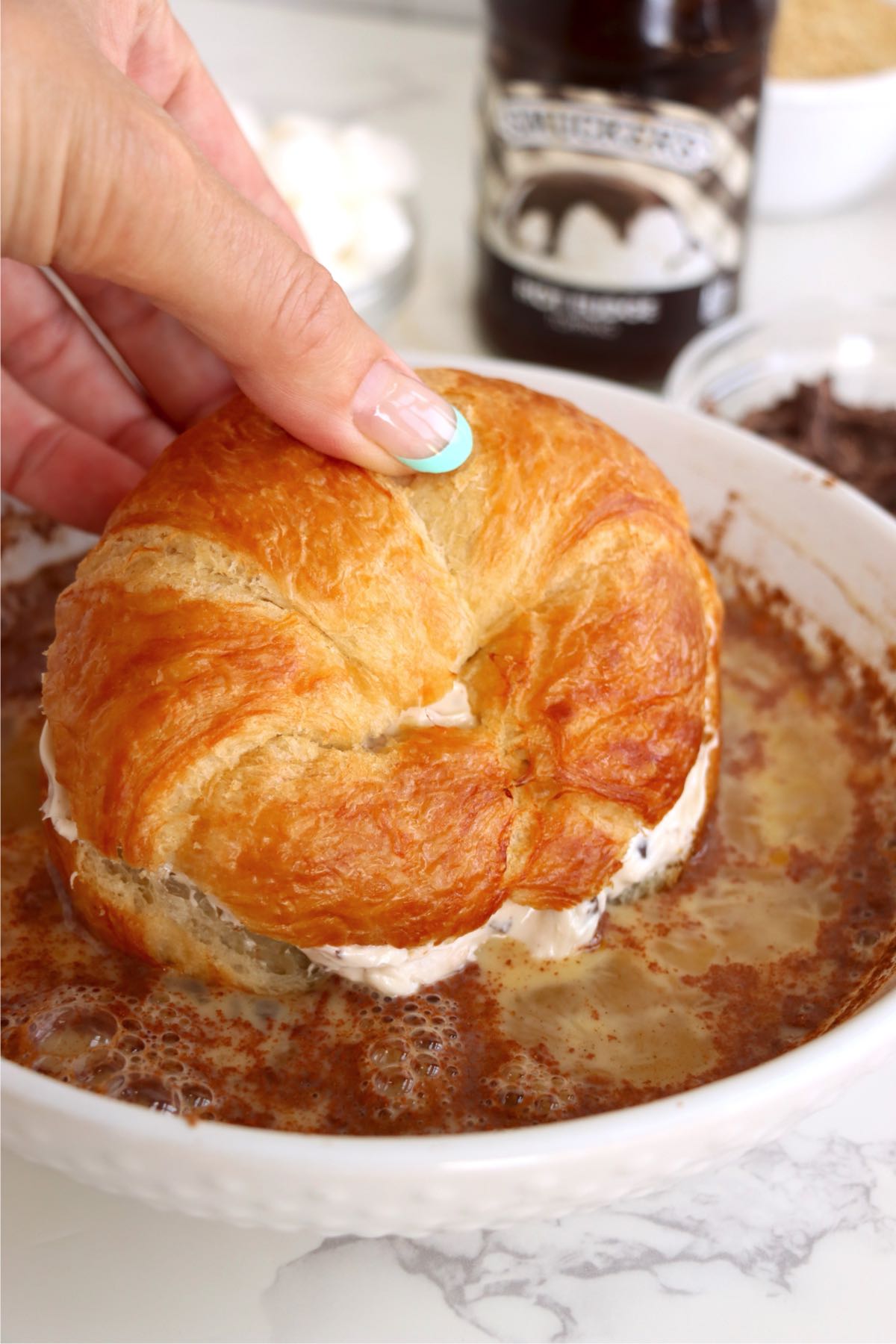 Hand lowering a cream-filled croissant into a cinnamon-speckled egg mixture in a shallow white bowl.