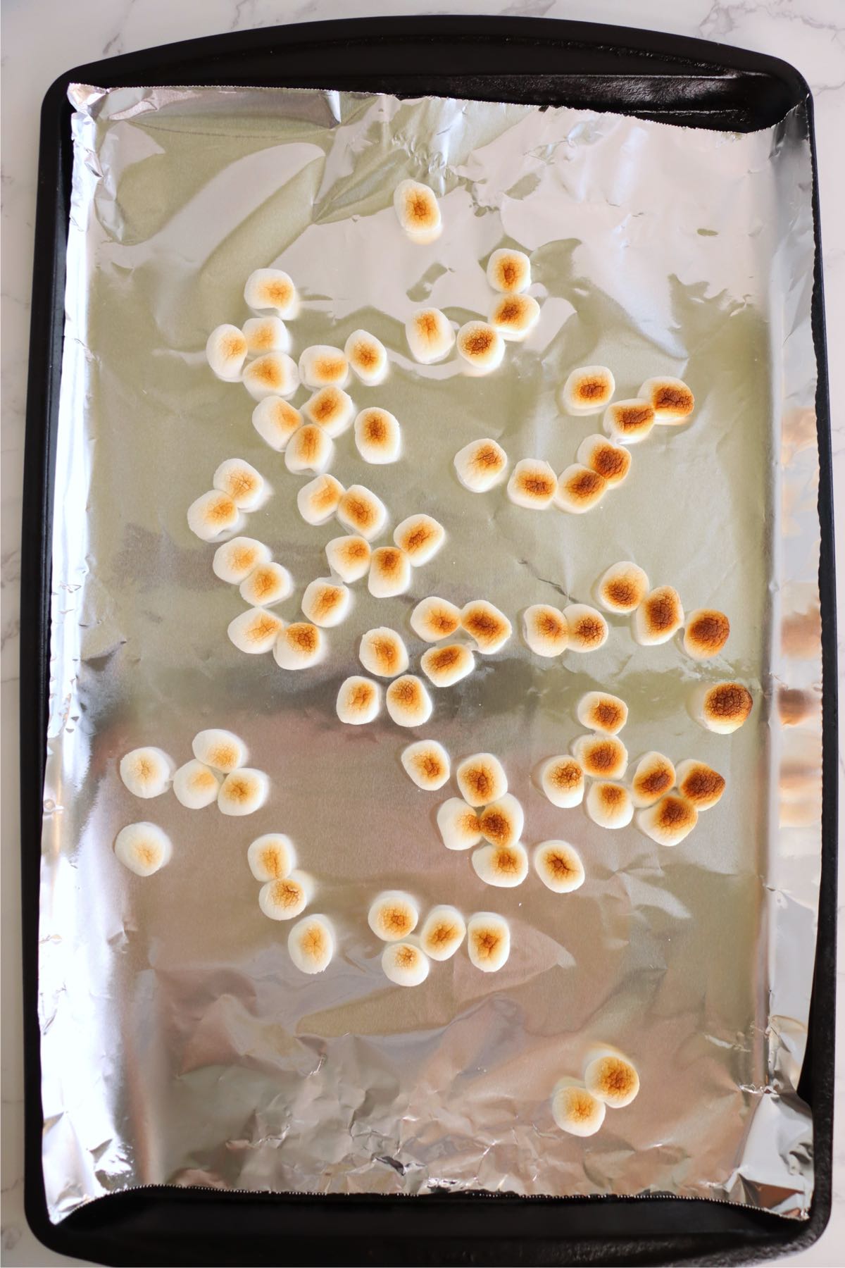 Mini marshmallows with toasted golden centers scattered across a foil-lined baking sheet.