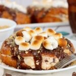 Stuffed croissant topped with chocolate sauce, graham crumbs and toasted marshmallows on a white plate beside a fork.