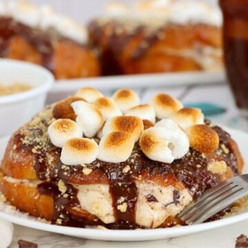 Stuffed croissant topped with chocolate sauce, graham crumbs and toasted marshmallows on a white plate beside a fork.