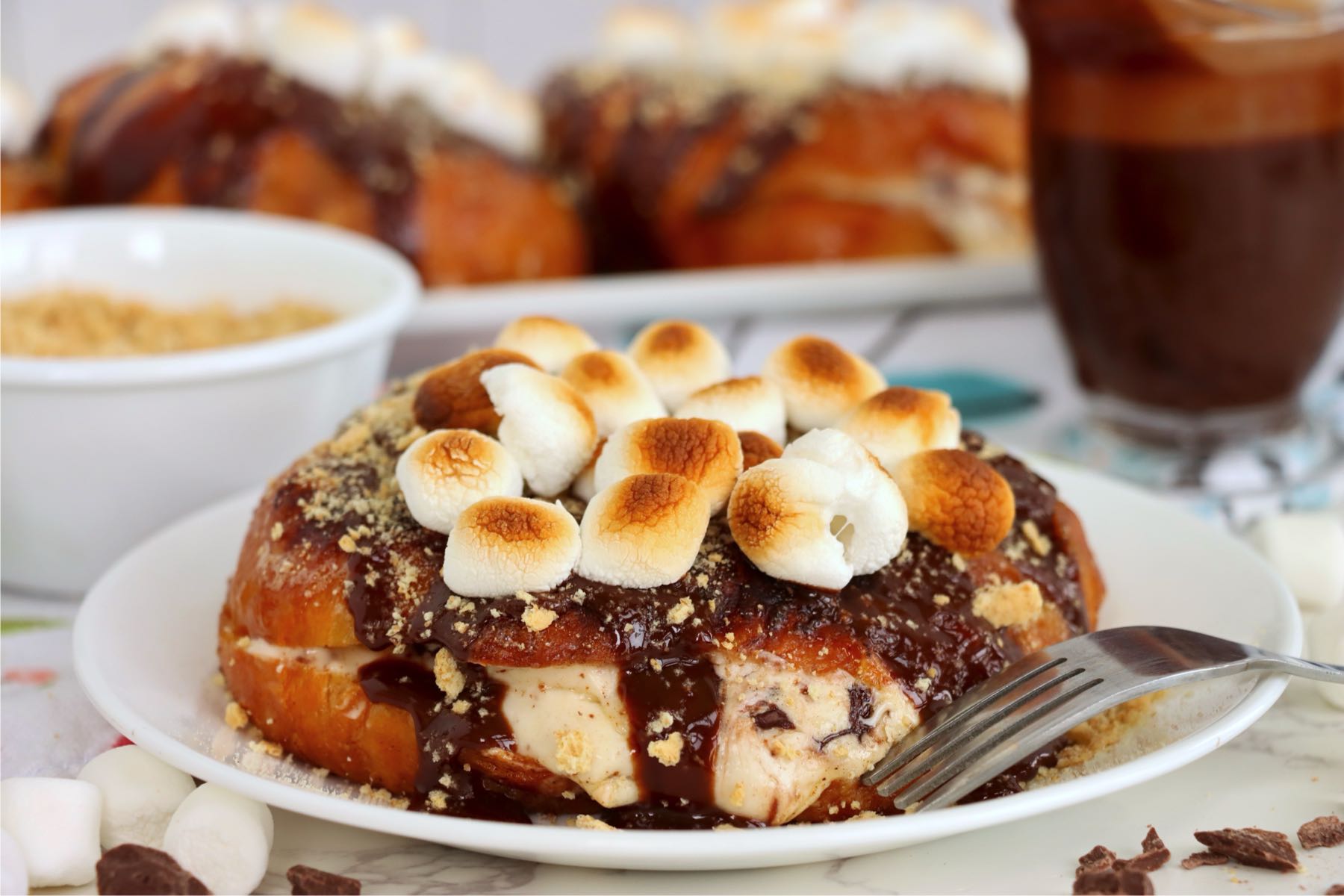 Stuffed croissant topped with chocolate sauce, graham crumbs and toasted marshmallows on a white plate beside a fork.