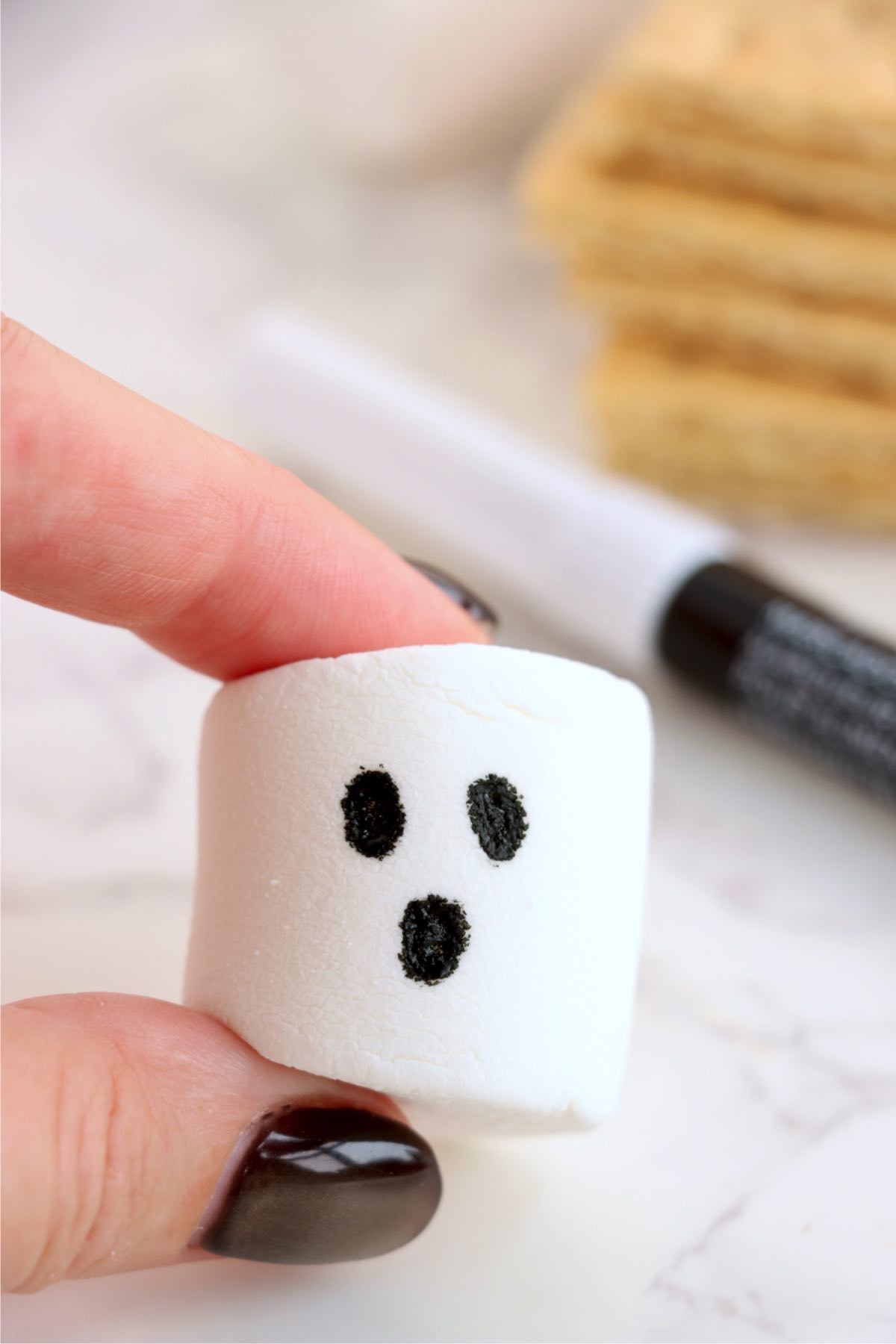 White marshmallow decorated with two black eyes and an oval mouth, held between fingers.