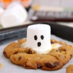 Baked chocolate chip cookie surrounding a white marshmallow ghost with black eyes and mouth.