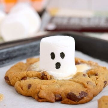 Baked chocolate chip cookie surrounding a white marshmallow ghost with black eyes and mouth.