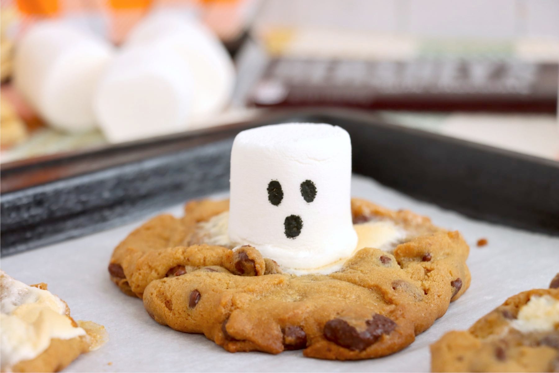 Baked chocolate chip cookie surrounding a white marshmallow ghost with black eyes and mouth.