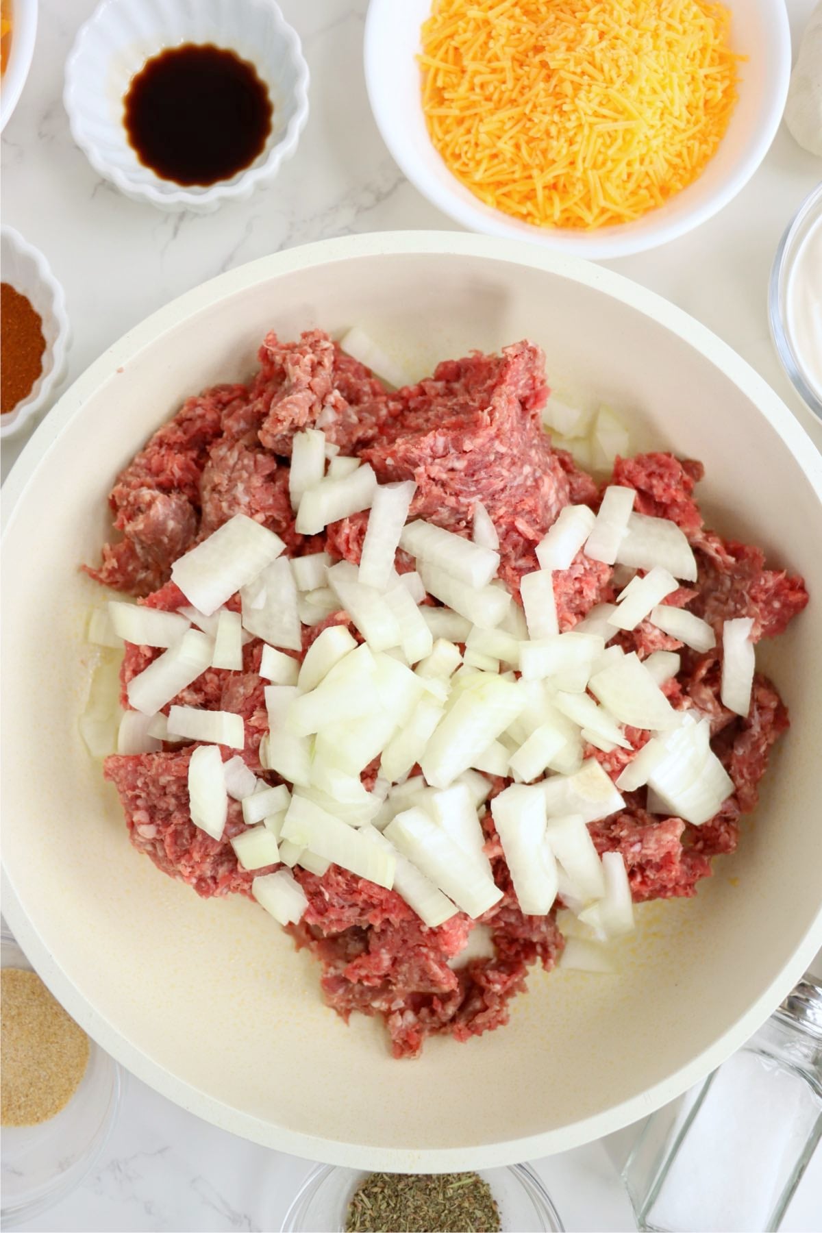 Raw ground beef and diced yellow onion added to a large skillet surrounded by seasonings and shredded cheese.