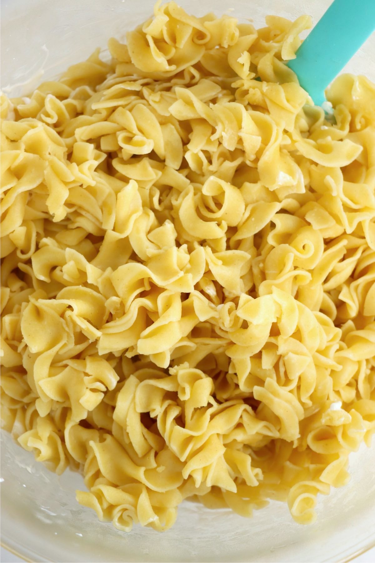 Cooked wide egg noodles piled in a clear mixing bowl beside a turquoise silicone spatula.